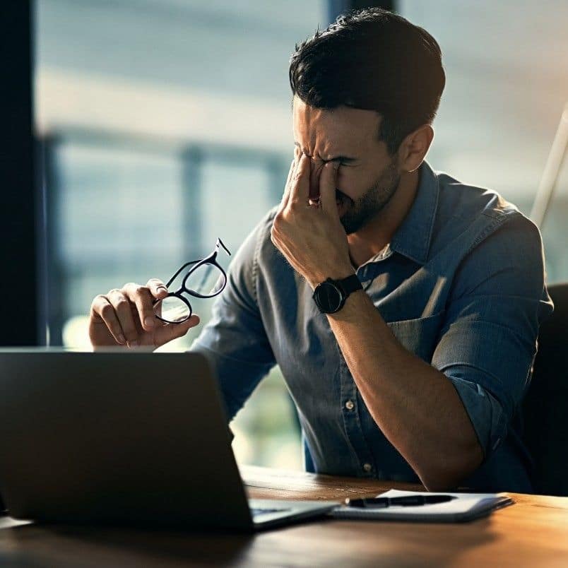 A man sitting at a desk with a laptop, holding his glasses in one hand and rubbing his eyes with the other, appears stressed—highlighting the need for emotional intelligence in project management within a dimly lit office setting.