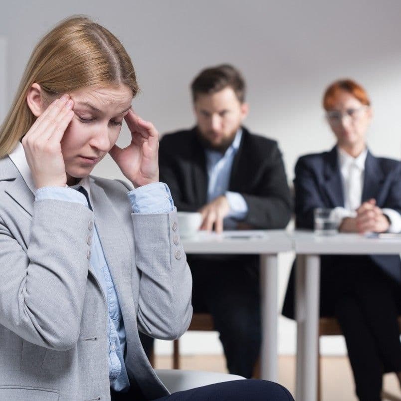 A woman in business attire, embodying leadership under pressure, sits holding her temples and looking stressed, while two people in suits observe her from a desk in the background.