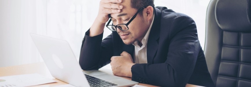 A man in a suit sits at a desk, looking stressed while holding his head with one hand and staring at his laptop screen during pmp exam prep.