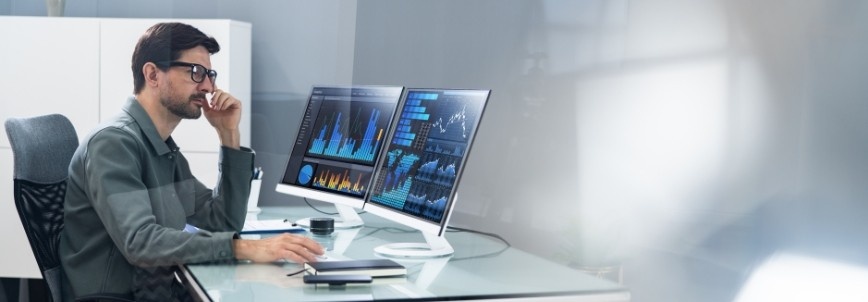 A man sits at a desk in an office, looking at two computer monitors displaying colorful data charts and graphs on a project dashboard. A keyboard, mouse, notebook, and pen are on the desk.