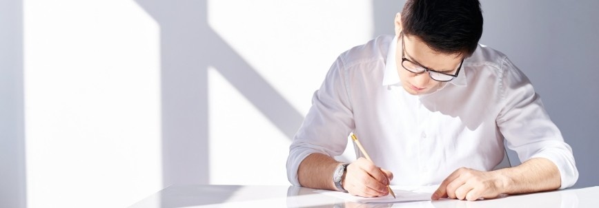 A certified associate in project management, wearing glasses and a white shirt, sits at a white desk, writing on a piece of paper. Bright sunlight creates shadows on the white wall behind him.