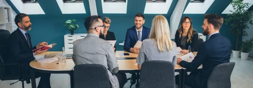 A group of eight professionals in business attire sit around a conference table in a modern office, engaged in a meeting and conversation within a dynamic project environment, with papers and devices on the table.