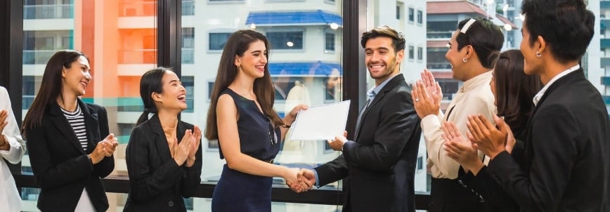 A woman receives her CAPM certification and a congratulatory handshake from a man in an office setting, surrounded by colleagues clapping and smiling in celebration.