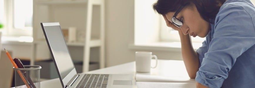 A person wearing glasses sits at a desk with a laptop, looking stressed or tired while studying how to get through CAPM. Papers, a pencil holder, and a mug are on the desk in a bright room.