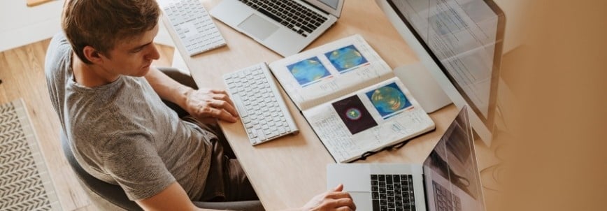 A person sits at a desk working on a laptop, with another monitor, keyboard, a notebook displaying colorful charts from their CAPM course, and a smartphone nearby. The workspace is organized and well-lit.