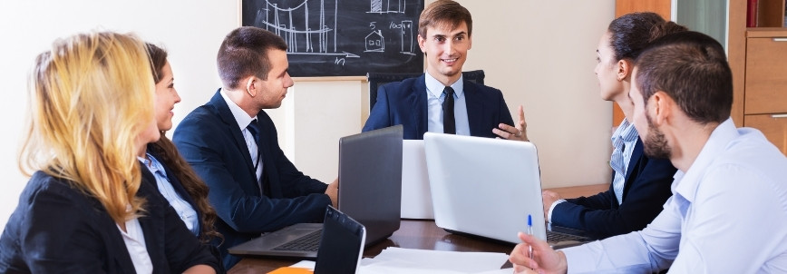 Five business professionals sit around a table with laptops, attentively listening to a man in a suit discussing PMP certification. A chalkboard with diagrams is visible in the background.