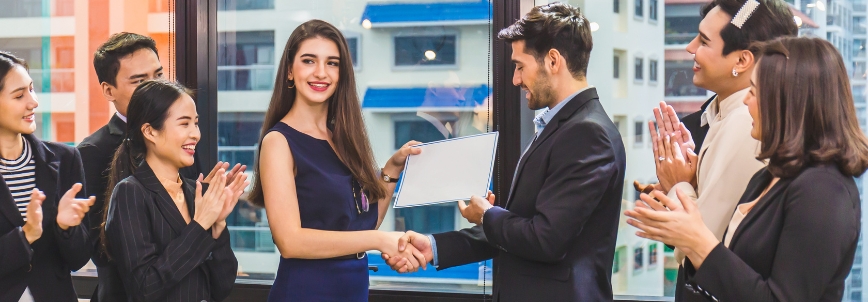 A woman in formal attire receives a certificate, celebrating the benefits of PMP certification, as she shakes hands with a man in an office while colleagues around them smile and applaud.