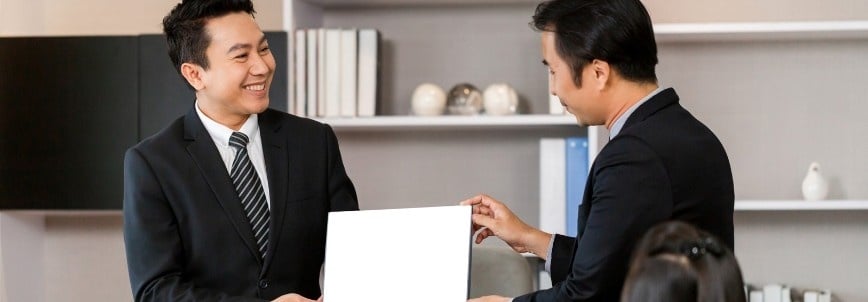Two men in business suits smiling and holding a blank white sheet of paper together in an office setting, perhaps discussing CAPM certification requirements, with shelves and decorative items in the background.