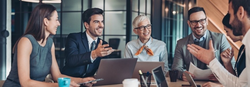A group of business professionals sit around a table in an office, smiling and having a lively discussion about PMP process groups. Laptops, documents, and coffee cups are on the table, with large glass windows in the background.