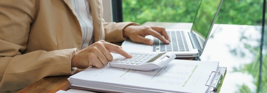 A person in a beige blazer uses a calculator and types on a laptop at a desk with financial documents and a clipboard, possibly researching PMP certification cost, with greenery visible outside the window.
