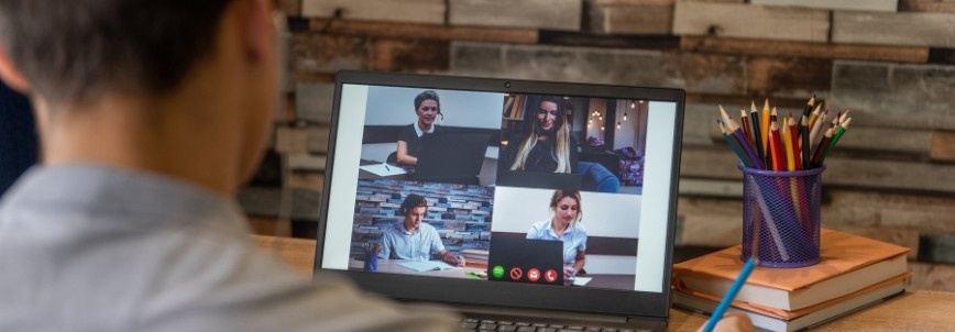 A person sits at a desk with a laptop, participating in an agile team collaboration video call with four people. A cup filled with colored pencils and stacked books are next to the laptop, reflecting a hybrid work model environment.