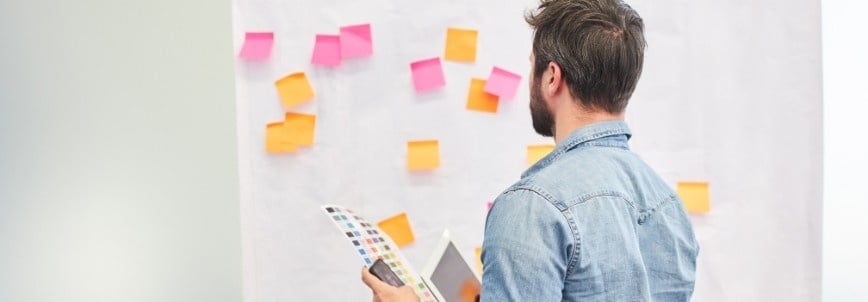 A man in a denim shirt stands in front of a whiteboard covered with colorful sticky notes, holding capm flashcards and a smartphone, looking at the notes.