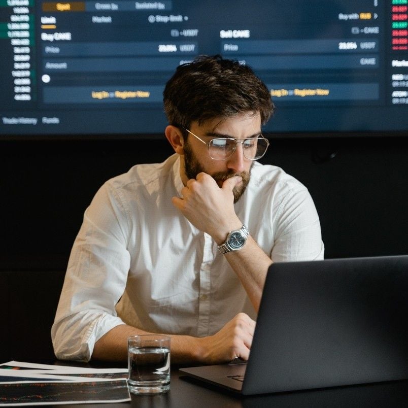A man with glasses and a beard sits at a desk in a project environment, looking thoughtfully at a laptop. A glass of water and papers are on the desk. A large screen displaying financial data is in the background.