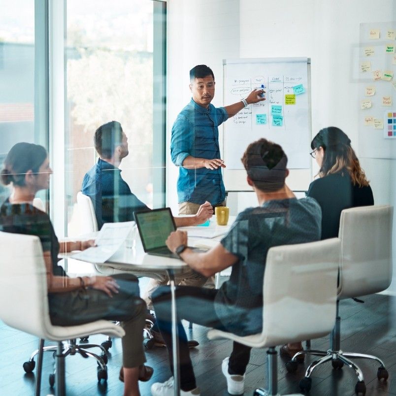 A man, a certified associate in project management, stands by a whiteboard with sticky notes, presenting to four colleagues seated around a conference table with laptops and papers in a modern office.
