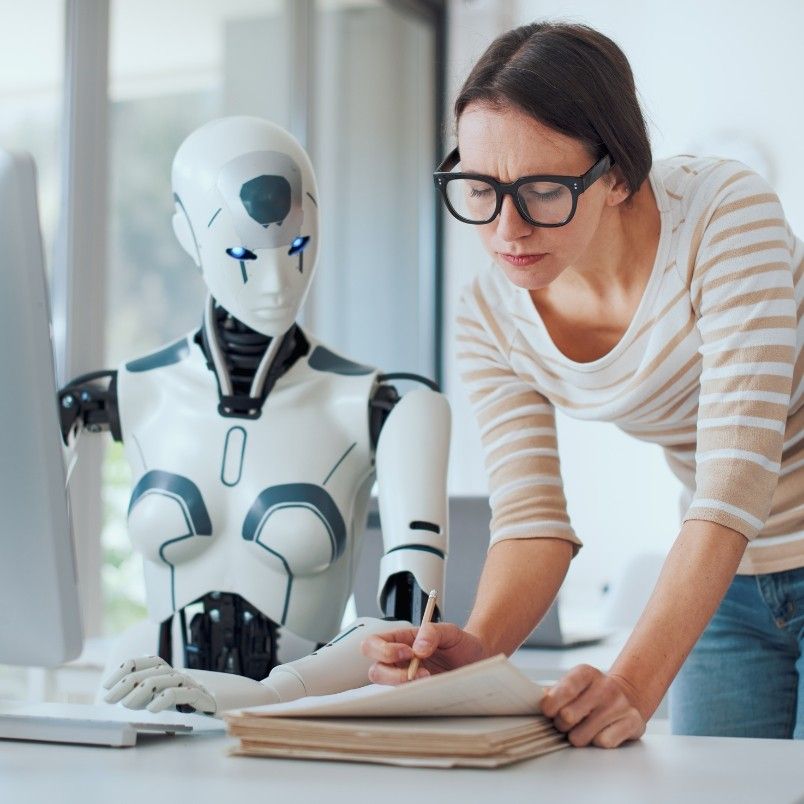 A woman wearing glasses writes on a notepad next to a humanoid robot at a desk, both looking at papers and a computer screen—an example of agile team collaboration in a hybrid work model in a bright office setting.