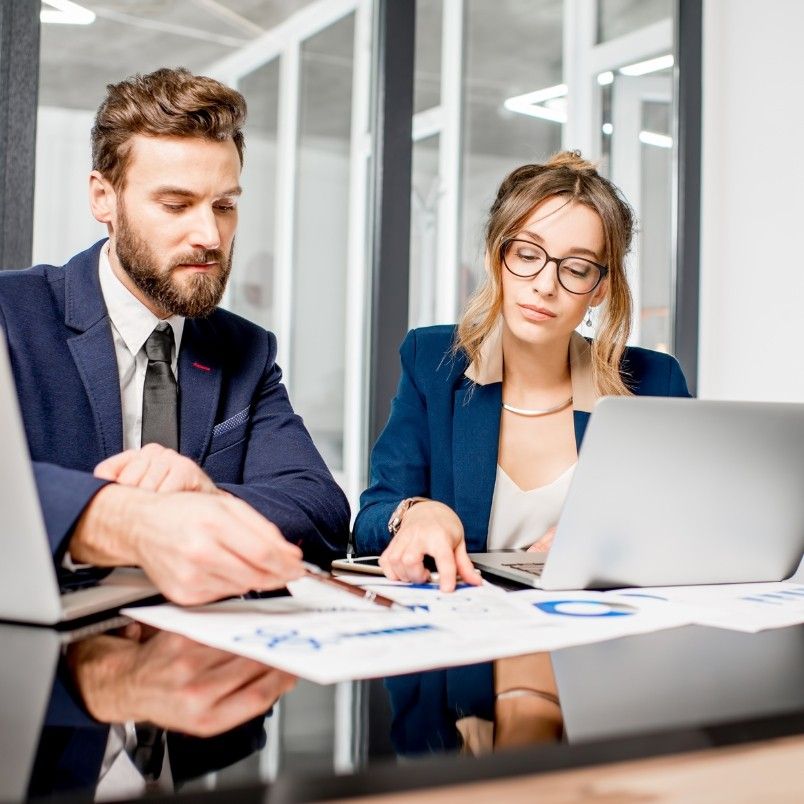 Two business professionals, a man and a woman, both with capm cert credentials, sit at a desk with laptops, reviewing documents and charts together in a modern office setting.