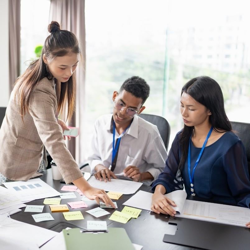 Three people sit at a table covered with papers and colorful sticky notes. One woman stands, pointing at a note, while the others listen. They appear to be collaborating on a project, possibly preparing for the CAPM cert exam.