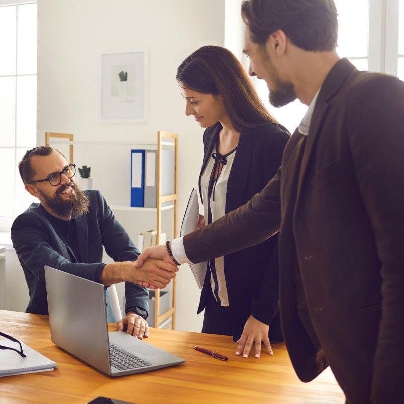 Three business professionals interact in a modern office. A seated man with a beard, a certified associate in project management, shakes hands with a standing man, while a woman stands beside them, smiling. A laptop and documents are on the table.