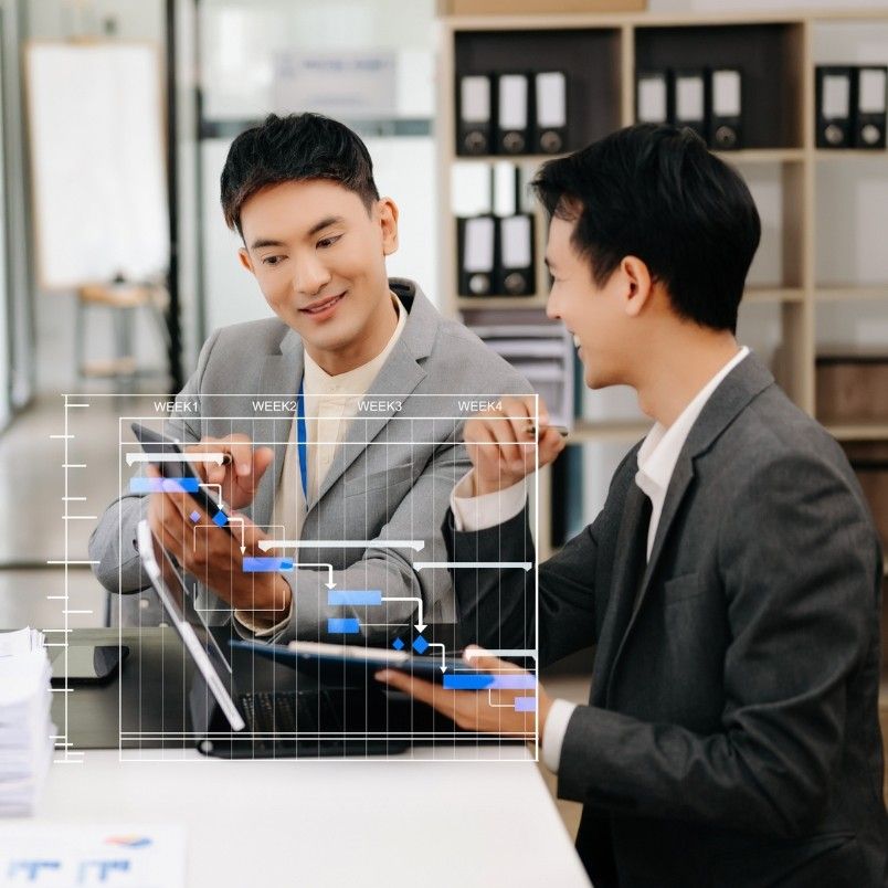 Two men in suits sit at a desk, discussing and pointing at a digital Gantt chart powered by generative AI in project management, highlighting advanced project planning and collaboration in a modern office setting.