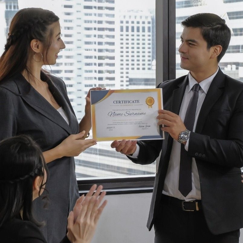 A man in a suit presents a certificate recognizing capm certification requirements to a smiling woman in business attire, while another seated woman claps. They are in an office with large windows and city buildings in the background.