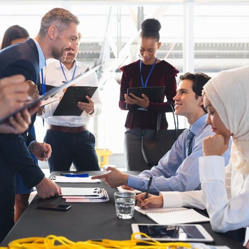 A diverse group of professionals interacts at a business meeting, discussing strategies on how to get through CAPM. Some stand and write on clipboards, while others sit at a table with documents, notepads, and glasses of water, engaged in conversation.