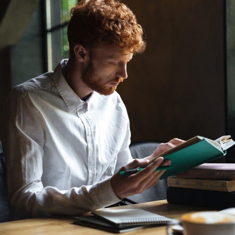 A young man with curly red hair and a white shirt sits at a table reading a book about the CAPM course, holding a pencil, with a notebook and a cup of coffee nearby in a softly lit room.