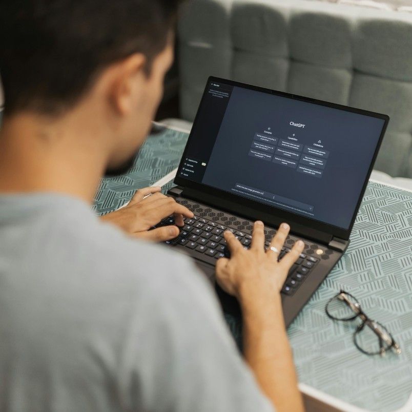A person types on a laptop displaying the ChatGPT interface, exploring generative AI in project management, while sitting at a table with a blue patterned tablecloth. A pair of eyeglasses rests beside the laptop.