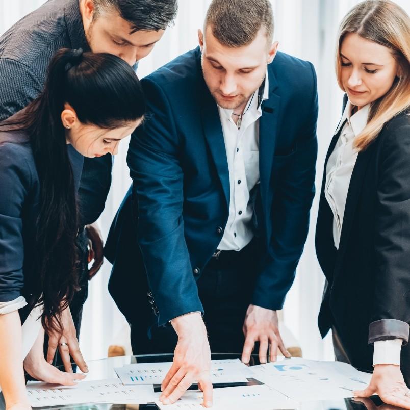 Four business professionals stand around a table, closely examining and discussing documents and charts, suggesting a collaborative meeting or brainstorming session for a CAPM course in a bright office environment.