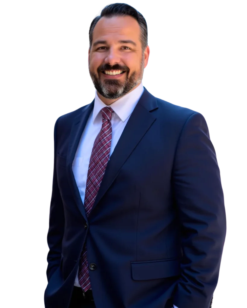 A man with dark hair and a beard, wearing a navy blue suit, white shirt, and red patterned tie, is smiling and standing with one hand in his pocket against a plain background.