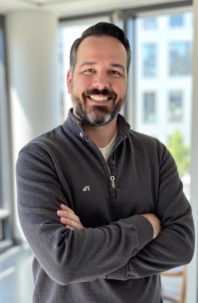 A man with a beard and mustache smiles with arms crossed, wearing a dark gray pullover in a brightly lit modern office with large windows.