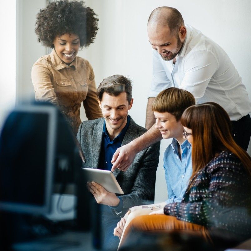 Five people gather around a tablet, smiling and collaborating in a bright office setting. Demonstrating agile team collaboration in a hybrid work model, one person points at the screen while others watch attentively. A computer monitor is visible in the foreground.