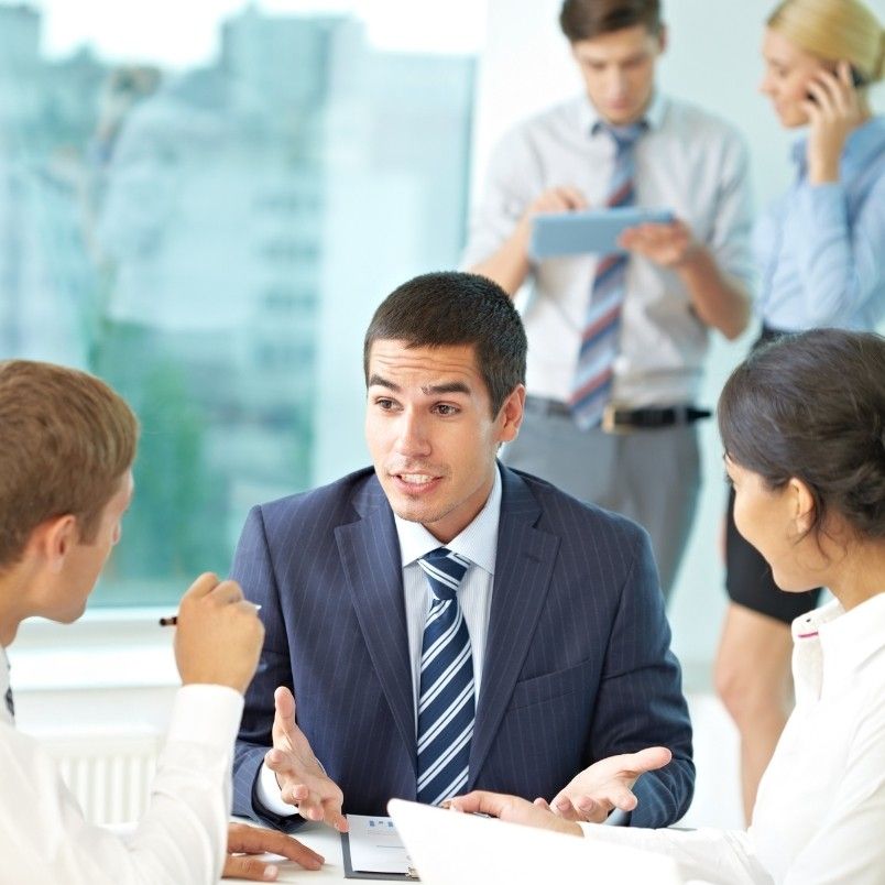 A man in a suit gestures while discussing project governance with two colleagues at a table. In the background, one person uses a tablet and another speaks on the phone in a modern office setting.