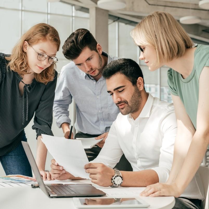 Four young professionals gather around a desk, looking at documents and a laptop screen, engaged in a focused project governance discussion in a bright, modern office setting.