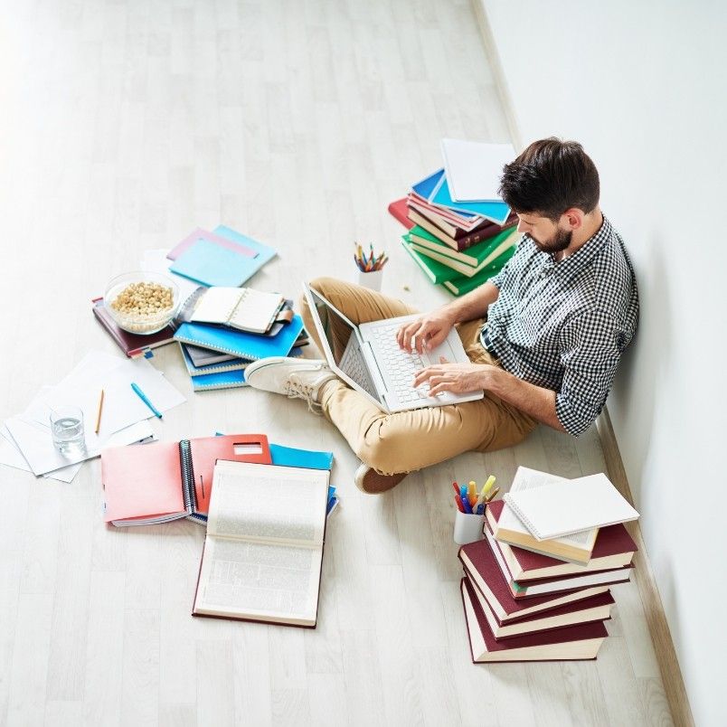 A man sits on the floor against a wall, typing on his laptop while preparing for his Certified Associate in Project Management exam, surrounded by piles of books, notebooks, folders, papers, a bowl of cereal, a glass of water, and stationery.