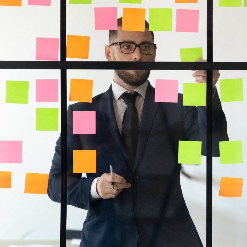 A man in a suit stands behind glass covered with colorful sticky notes, holding a pen and placing a note, appearing focused and thoughtful—perfectly capturing the intensity of pmp exam prep.