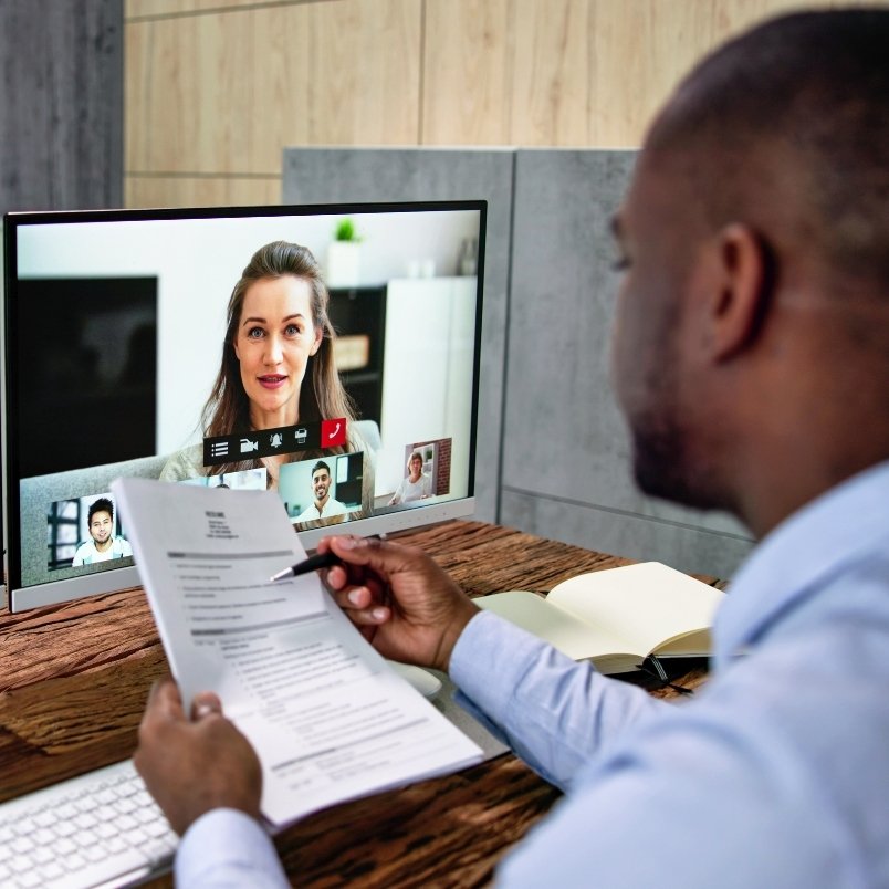 A man holding a resume sits at a desk, participating in a video call job interview for a position requiring CAPM certification, with several people visible on his computer screen. A notebook and pen are also on the desk.
