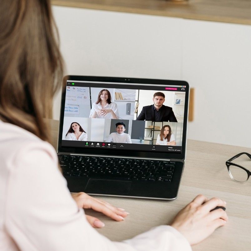 A person sits at a desk using a laptop for a video conference with four others, each in separate windows—showcasing agile team collaboration in a hybrid work model. Glasses rest on the tabletop next to the laptop.