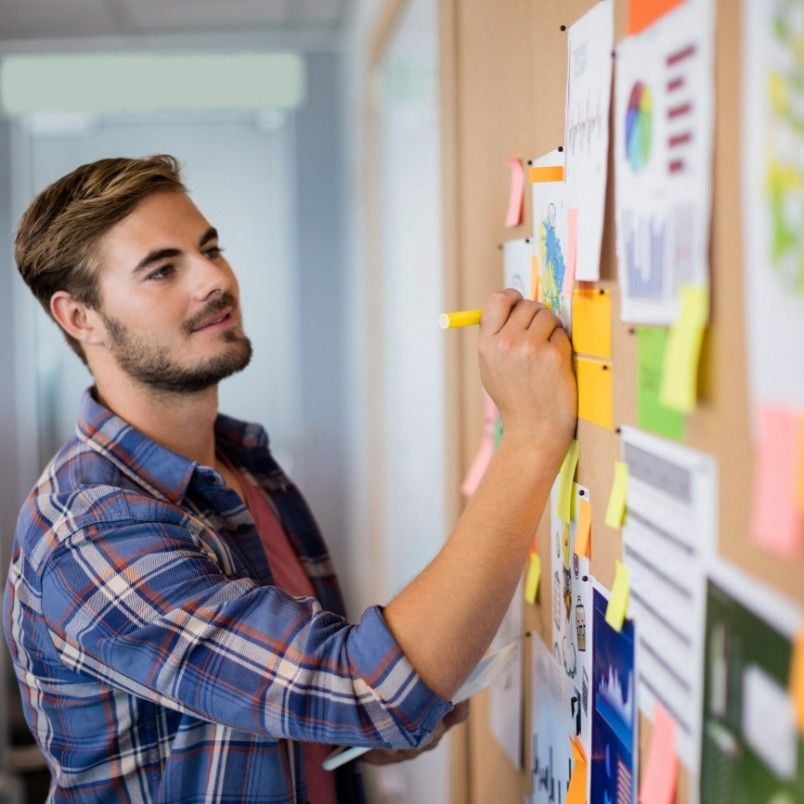 A man in a plaid shirt stands beside a corkboard covered with charts, graphs, capm flashcards, and sticky notes, writing on one of the papers with a yellow marker in a bright office setting.