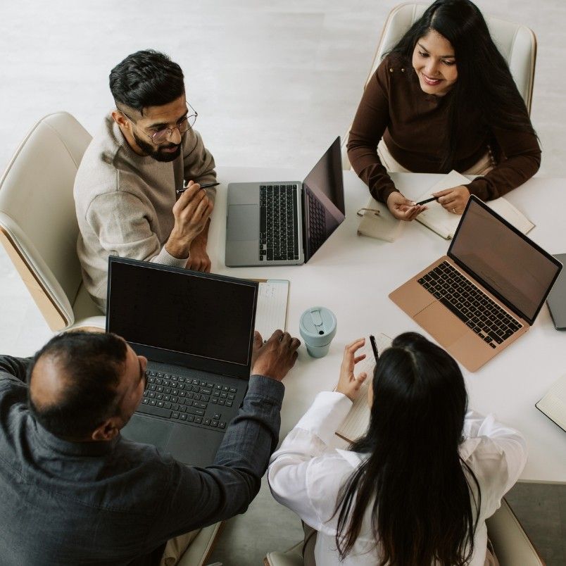 Four people sit around a table with laptops and notebooks, engaged in discussion. Two women and two men appear to be collaborating as an agile team in a modern, bright office that supports a hybrid work model.