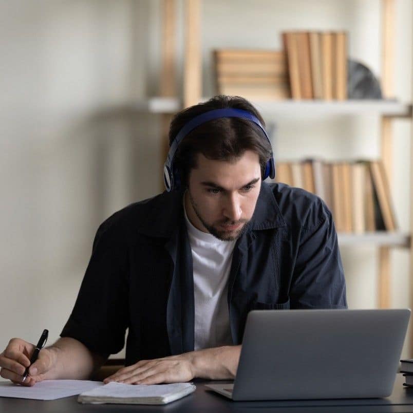 A young man wearing headphones sits at a desk, focused on his laptop as he documents his PMP Exam Experience. He writes notes in a notebook, with bookshelves and books in the background.