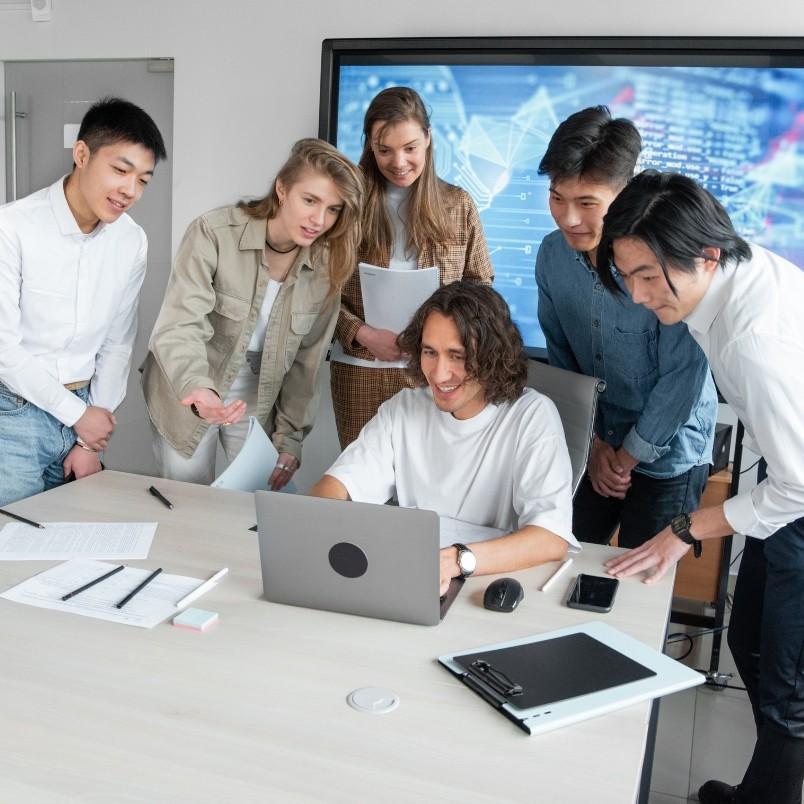 Six young adults gather around a table, smiling and looking at a laptop screen. Papers, charts, and a smartphone are scattered about as they discuss how generative AI in project management can enhance their workflow; digital graphs display in the background.