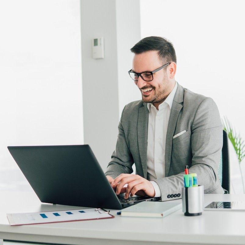 A man in a gray suit and glasses is smiling while working on a laptop at a white desk, exploring generative AI in project management, with office supplies and documents nearby in a bright, modern office.