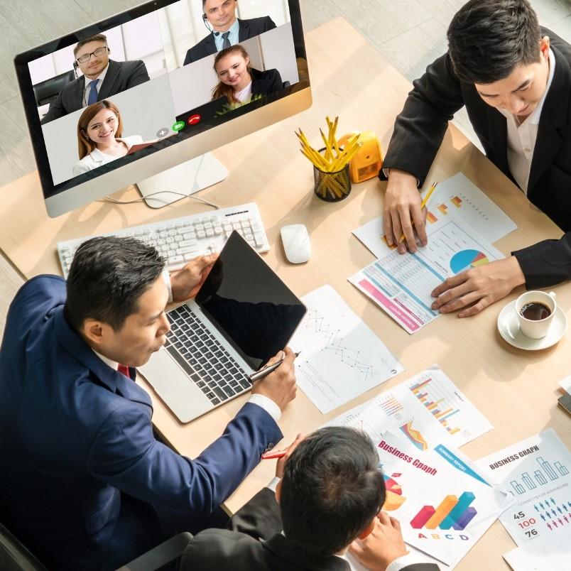Three people in suits sit at a desk with charts and documents, joined by four colleagues on a video call displayed on a monitor, demonstrating agile team collaboration in a hybrid work model for business growth analysis.