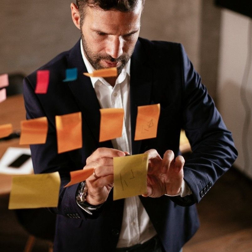 A man in a suit writes on a yellow sticky note while standing in front of a glass wall covered with colorful capm flashcards and sticky notes in an office setting.