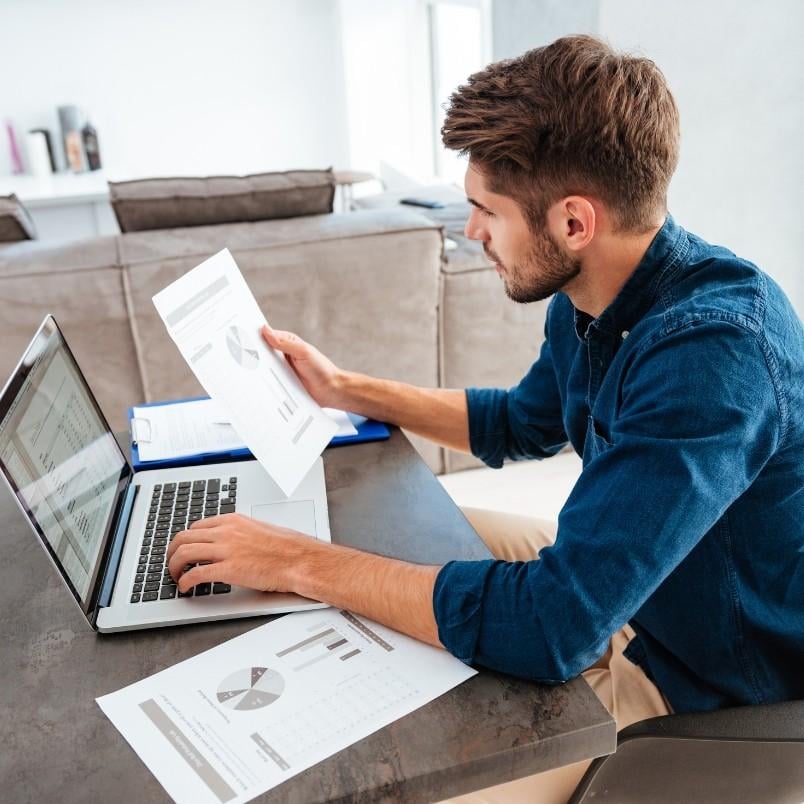 A man sits at a desk working on a laptop, holding a paper with charts and graphs. CAPM flashcards and more documents with charts are spread out on the table in front of him.