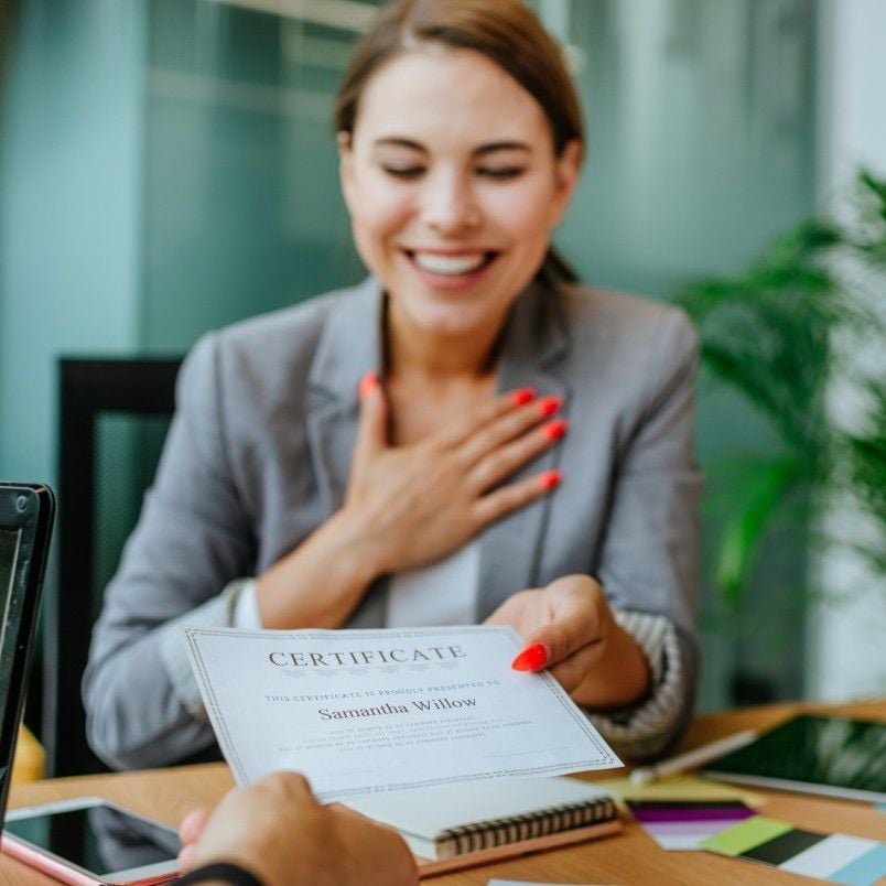 A smiling woman in a gray blazer holds a certificate with one hand and touches her chest with the other, looking happy and appreciative in an office setting after meeting CAPM certification requirements.