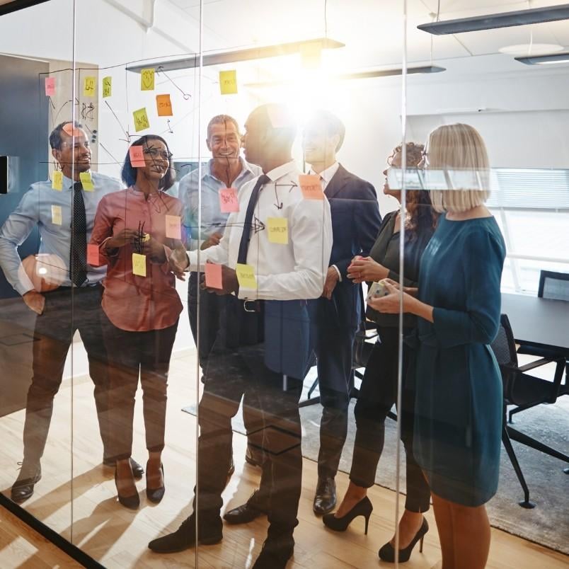 A diverse group of professionals stands in a glass-walled office, smiling and discussing colorful sticky notes and CAPM flashcards on the wall, suggesting a collaborative brainstorming or planning session.