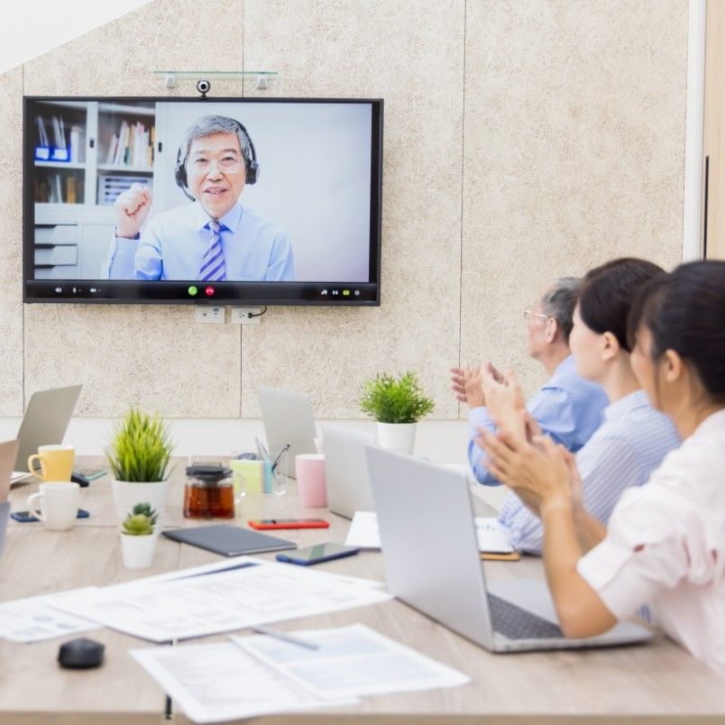 A group of people sitting at a conference table with laptops applaud a man wearing headphones, who is speaking on a video call displayed on a large screen—showcasing agile team collaboration in a hybrid work model in a modern office.