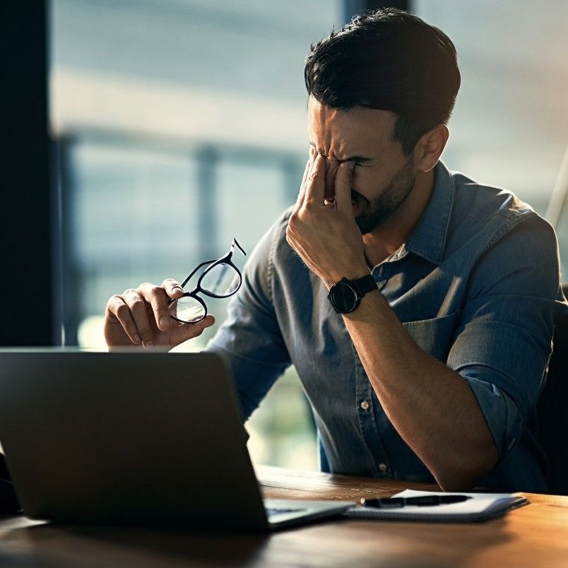 A man sitting at a desk in front of a laptop holds his glasses in one hand and rubs his eyes with the other, looking stressed or fatigued in a dimly lit office—perhaps struggling to keep up without generative AI in project management.