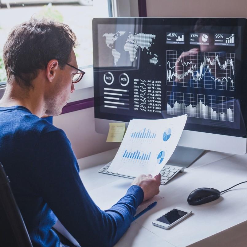 A man sits at a desk analyzing printed charts and graphs, while reviewing financial data and analytics on a computer screen displaying a project dashboard. A smartphone, mouse, and keyboard are on the desk.